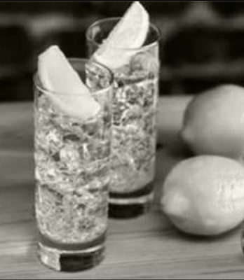 Black and white image of two tall cocktail glasses on table with lemon slices in each and whole lemons on table